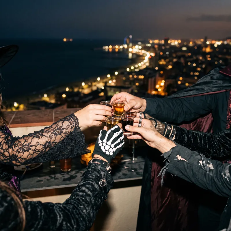 Costumed hands, one in a skeleton glove, clinking shot glasses above the Barcelona coastline